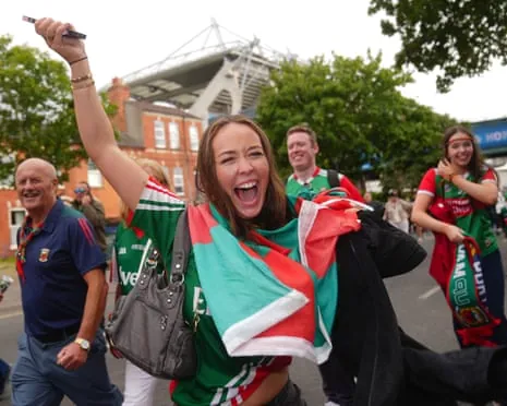 A woman wearing a green and red striped jersey and flag raises her arm and cheers outside a stadium